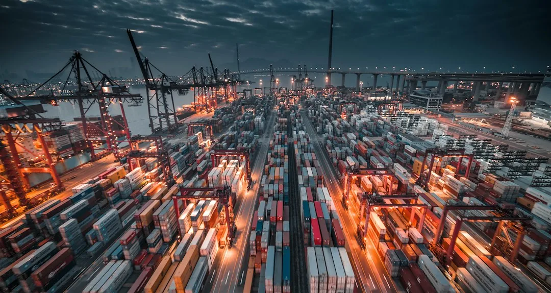 aranceles China México: A busy shipping port with stacked containers and cranes under a dark cloudy sky